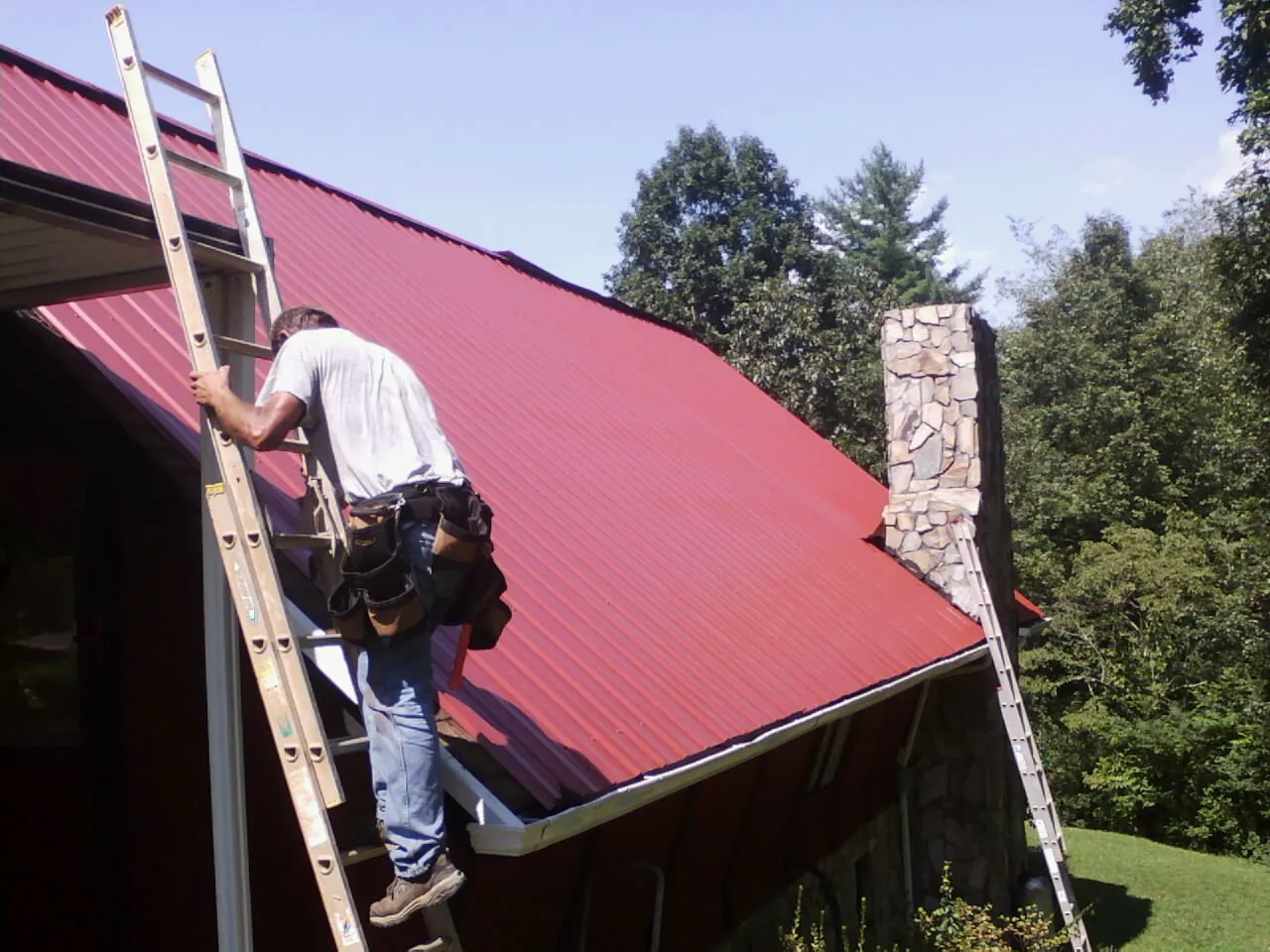 Roofer on a ladder beside a red metal roof and stone chimney — Soffit & Fascia Repair by Roofing Brothers in Valyermo